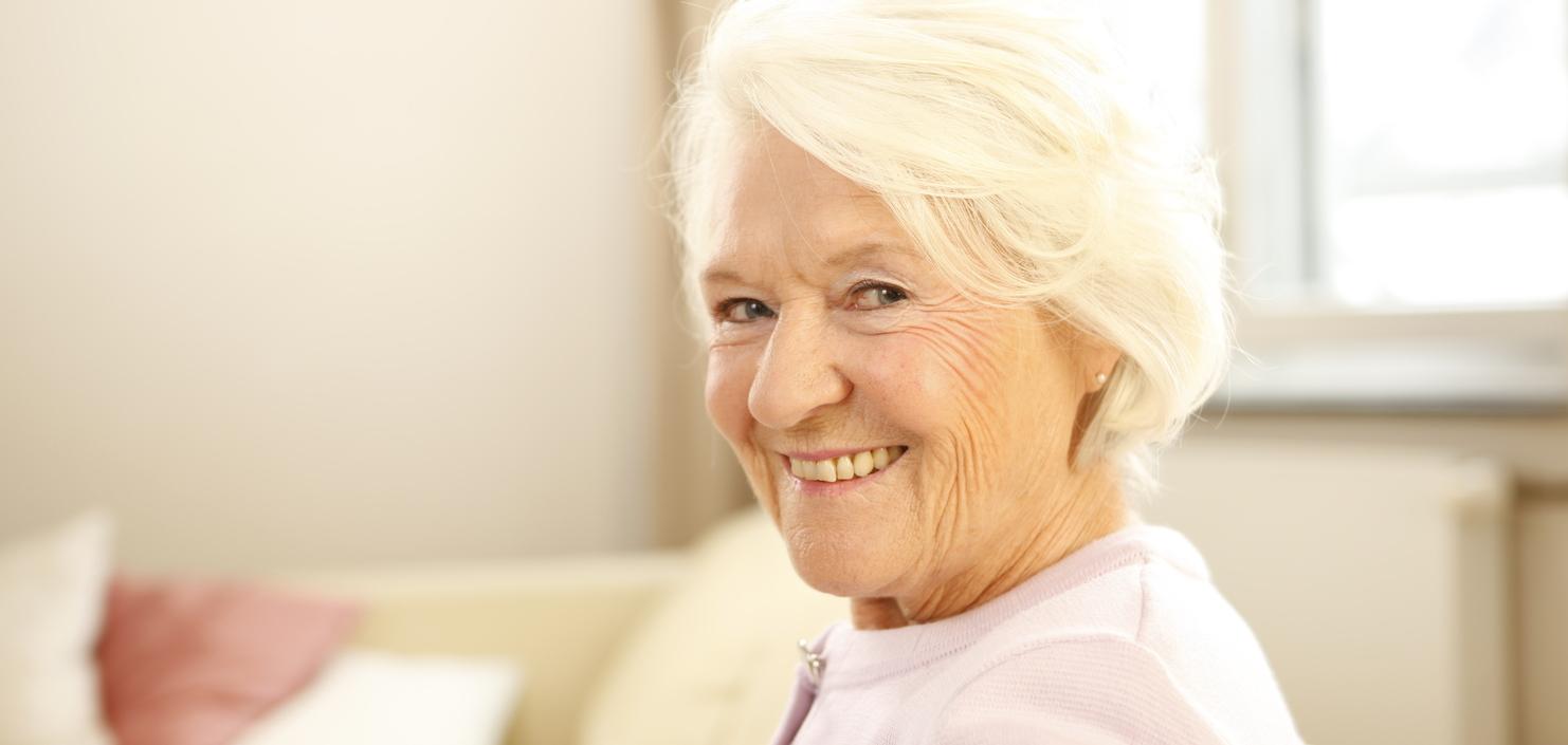 Lächelnde ältere Frau mit grauen Haaren sitzt auf einem Sofa in einem hellen Raum.